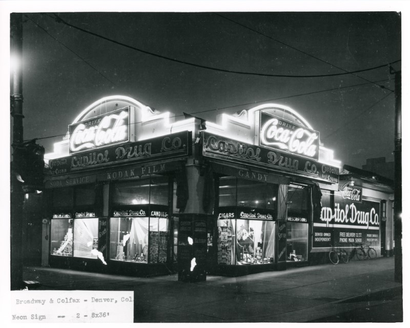 Nighttime photo of Capitol Drug Co., Broadway & Colfax 1930s. Predominant feature is a set of two identical large neon "Coca Cola" signs at the corner atop the one-story building.
