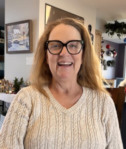 Photo of Carol Hamer, wearing a white sweater. Smiling, with shoulder-length auburn hair. 